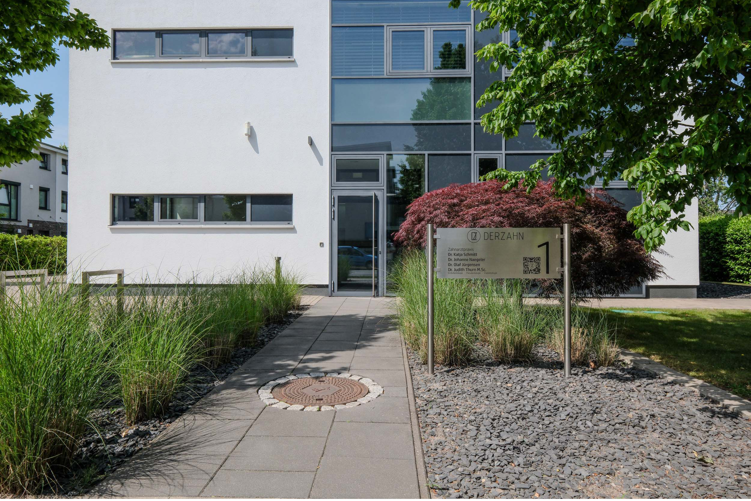 Außenansicht des Praxisgebäudes der Zahnarztpraxis "DERZAHN" in Oberneuland. Das Gebäude ist links weiß verputzt, mit großen Glasfronten im Eingangsbereich. Ein gepflasterter Zugangsweg ist rechts und links von Ziergräsern flankiert. Im Rechten Beet steht ein japanischer Ahorn und das Praxisschild mit den Namen der Behandelnden Zahnärzte und Zahnärztinnen. Dr. Katja Schmitt, Dr. Johanne Naegeler, Dr. Olaf Jürgensen und Dr. Judith Thurn M.Sc..